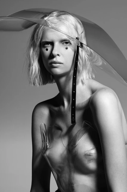 Monochrome portrait of a fair-skinned woman with blond hair and black dots on her face, wearing a transparent plastic top and headpiece.