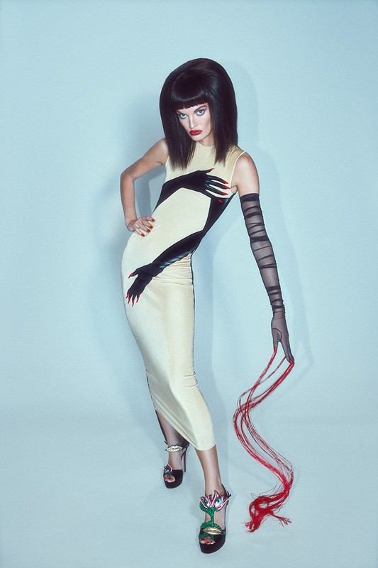 A woman poses in a cream dress with black hand designs, complemented by long black gloves and floral heels, against a light blue backdrop.