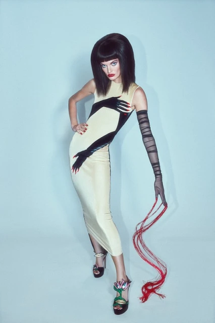 A woman poses in a cream dress with black hand designs, complemented by long black gloves and floral heels, against a light blue backdrop.