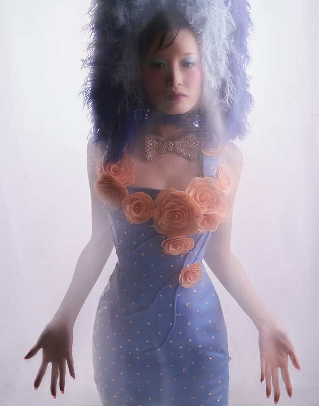 An Asian woman stands with her arms outstretched, wearing a blue dress decorated with orange rose appliques, a bow, and a large feathered headdress.
