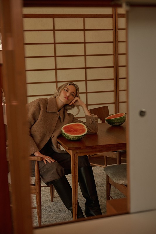 Woman in brown coat sits at table with watermelon halves indoors, near Japanese-style sliding doors, reflected in a mirror.