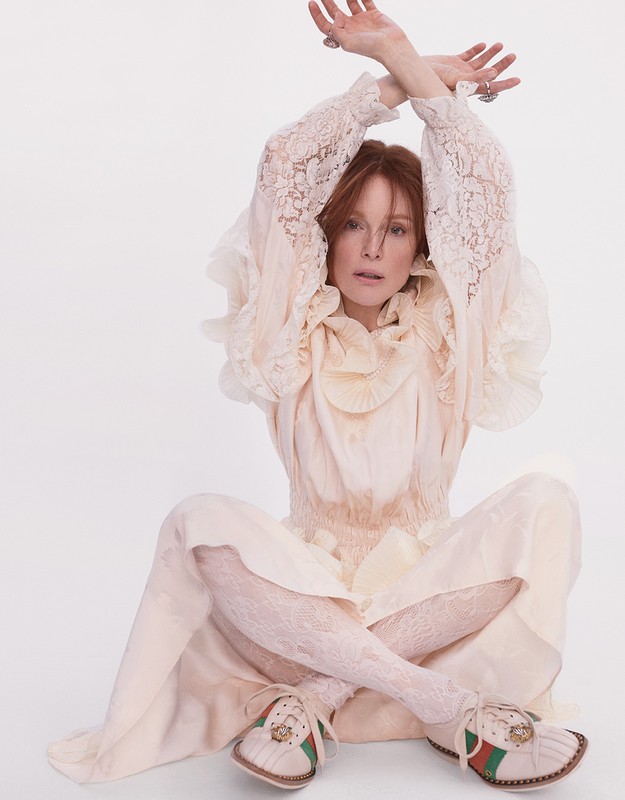 Woman with red hair poses in a white dress and lace tights sitting cross legged in front of a white background in a studio shot.