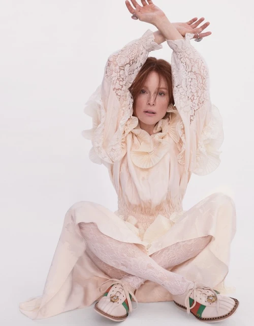 Woman with red hair poses in a white dress and lace tights sitting cross legged in front of a white background in a studio shot.
