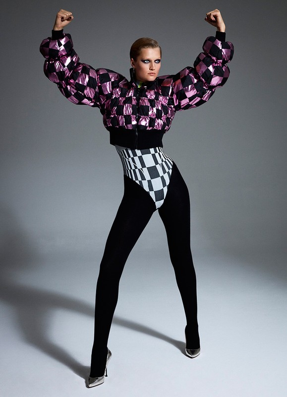 Full-length shot of a woman in a pink and black checkerboard bomber jacket, high-cut leotard, black tights, and metallic heels posing in a studio.