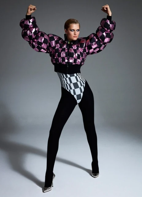 Full-length shot of a woman in a pink and black checkerboard bomber jacket, high-cut leotard, black tights, and metallic heels posing in a studio.