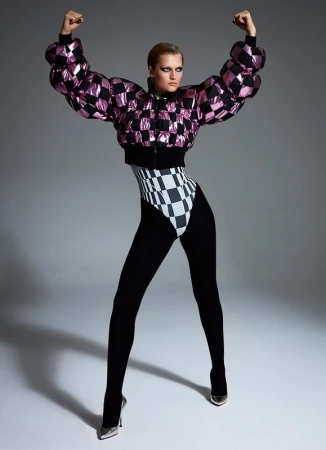 Full-length shot of a woman in a pink and black checkerboard bomber jacket, high-cut leotard, black tights, and metallic heels posing in a studio.