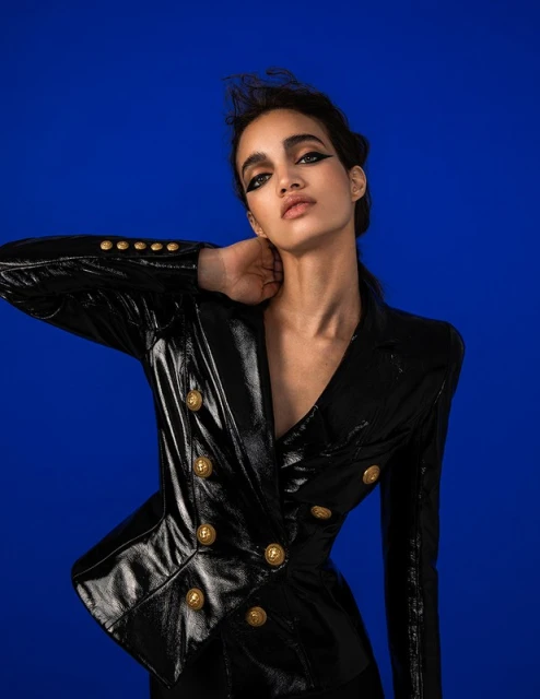 Portrait of a stylish woman in a black, shiny jacket with gold buttons against a solid blue background in a studio setting.
