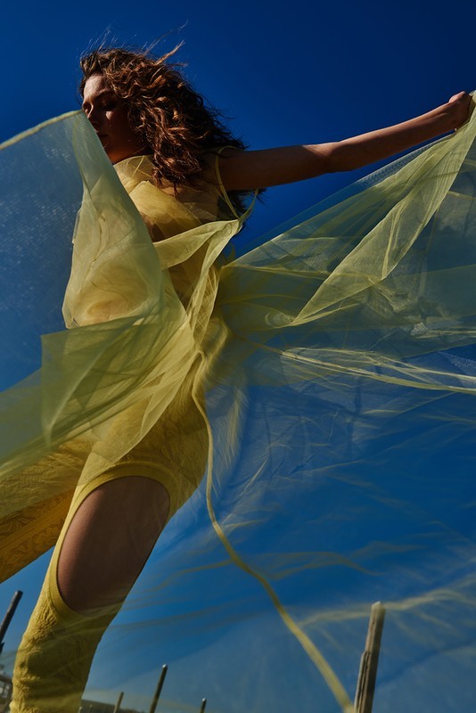 A woman in a yellow outfit poses outdoors against a blue sky, holding a sheer yellow fabric that floats around her in a low-angle fashion shot.