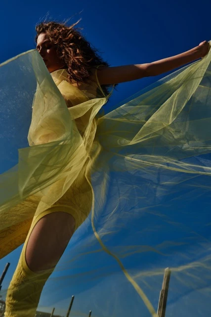 A woman in a yellow outfit poses outdoors against a blue sky, holding a sheer yellow fabric that floats around her in a low-angle fashion shot.