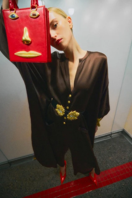 Woman poses indoors with a red purse featuring a face design, wearing a brown dress with gold embellishments and red heels.