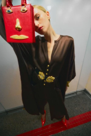 Woman poses indoors with a red purse featuring a face design, wearing a brown dress with gold embellishments and red heels.