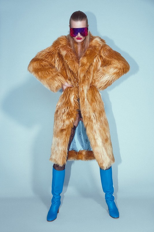 A blonde woman in an orange fur coat and blue boots poses against a light blue backdrop, accented by red lipstick and futuristic sunglasses.