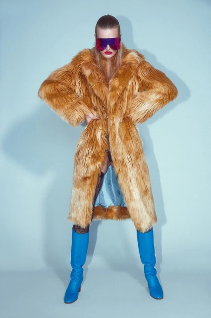 A blonde woman in an orange fur coat and blue boots poses against a light blue backdrop, accented by red lipstick and futuristic sunglasses.