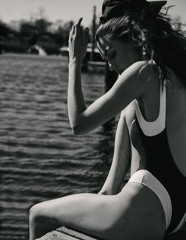 Monochrome portrait of a woman in a swimsuit sitting on a pier with her head in her hand against a blurred water background.