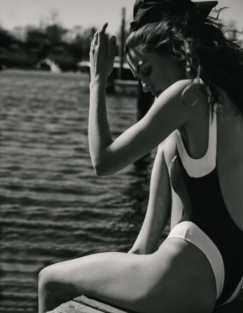 Monochrome portrait of a woman in a swimsuit sitting on a pier with her head in her hand against a blurred water background.