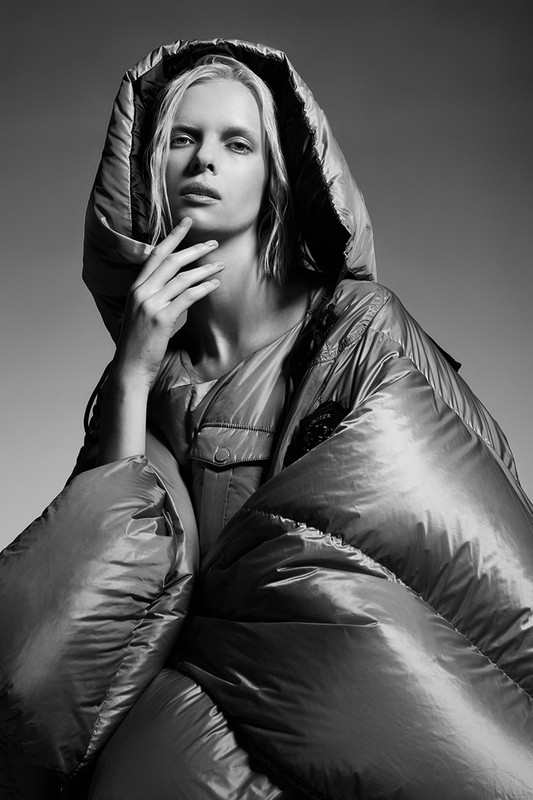Monochrome portrait of a blonde adult fashion model wearing an oversized hooded puffer coat and posing in a photo studio with hand to face.