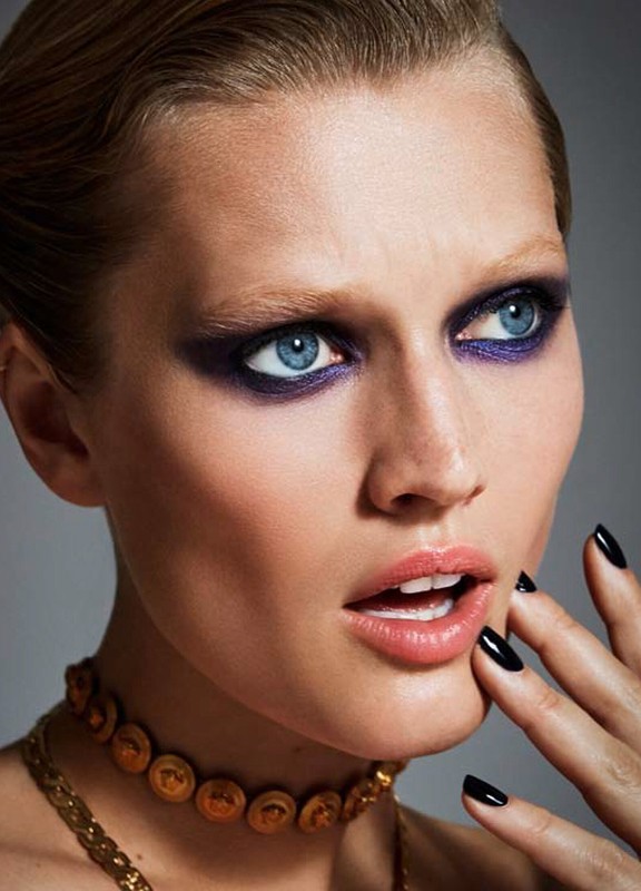 Close-up portrait of a woman with blue eyes, dark purple eye makeup, slicked-back hair, a gold choker, and black nail polish.