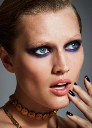 Close-up portrait of a woman with blue eyes, dark purple eye makeup, slicked-back hair, a gold choker, and black nail polish.