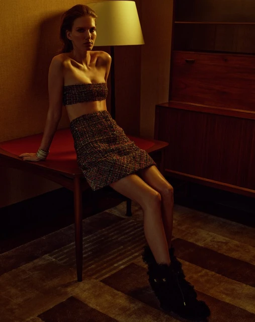 A woman wearing a brown tweed skirt and tube top set poses sitting on a table in a dimly lit room with mid-century modern furniture.