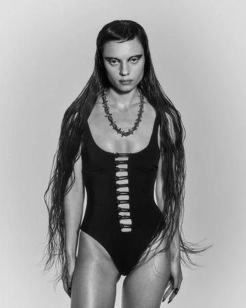 Monochrome portrait of a young woman with dark hair and dramatic eye makeup, wearing a black swimsuit and barbed wire necklace in studio.