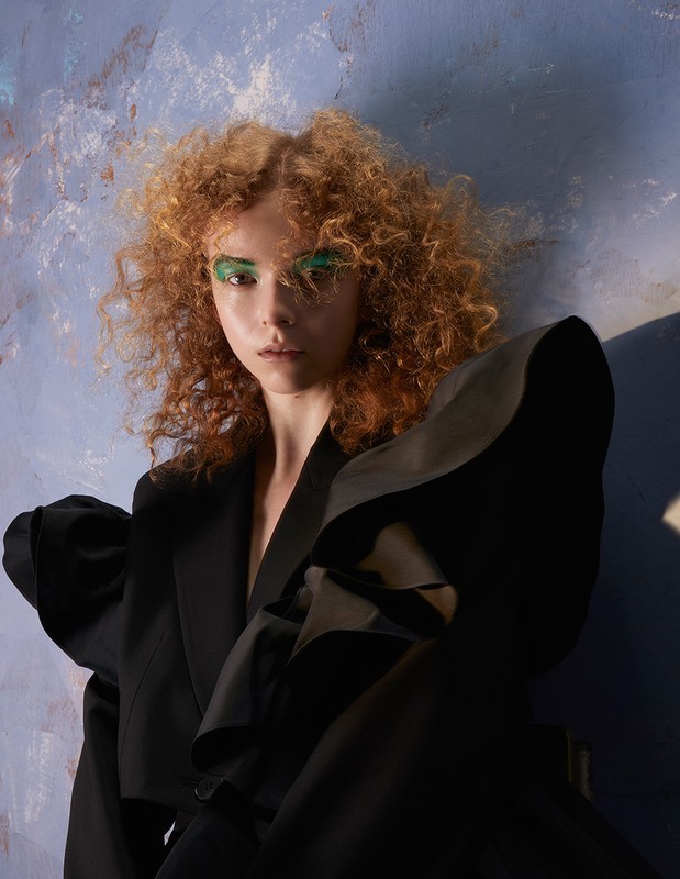Woman with curly red hair poses in dramatic black blazer with puffed sleeves against a textured blue studio background.