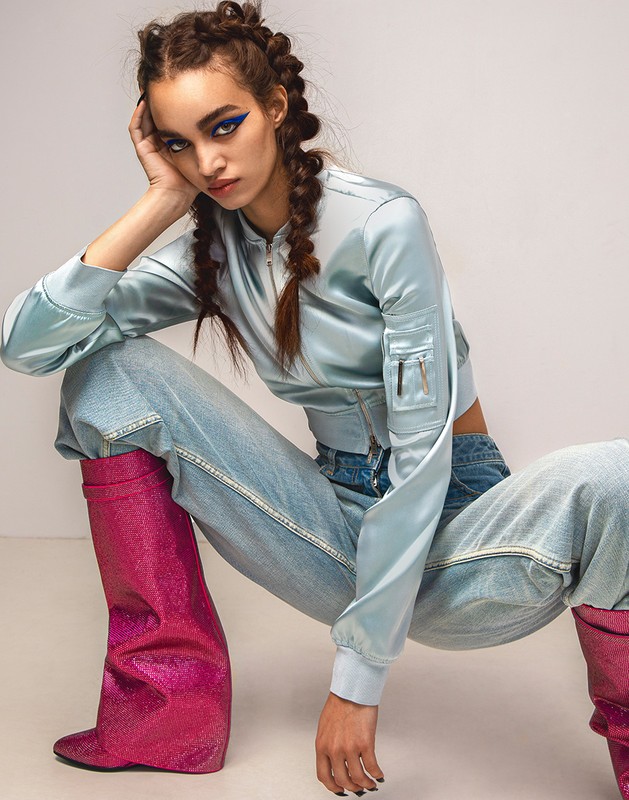 Model with braided hair and blue winged eyeliner wearing a light blue satin bomber jacket and pink glitter boots.
