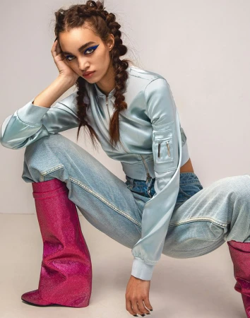 Model with braided hair and blue winged eyeliner wearing a light blue satin bomber jacket and pink glitter boots.