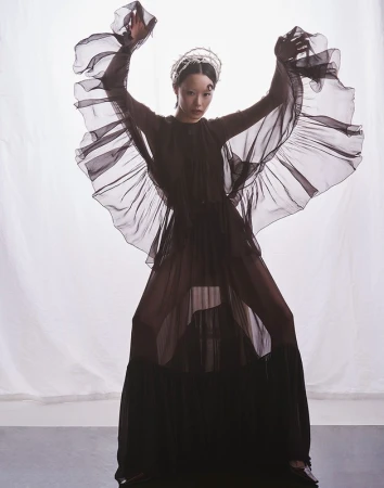 Model in a black sheer ruffled dress and tiara posing against a white background