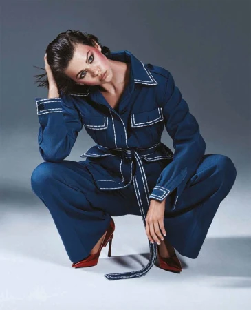 Woman wearing a blue belted suit with white contrast stitching and red heels squatting against a grey backdrop