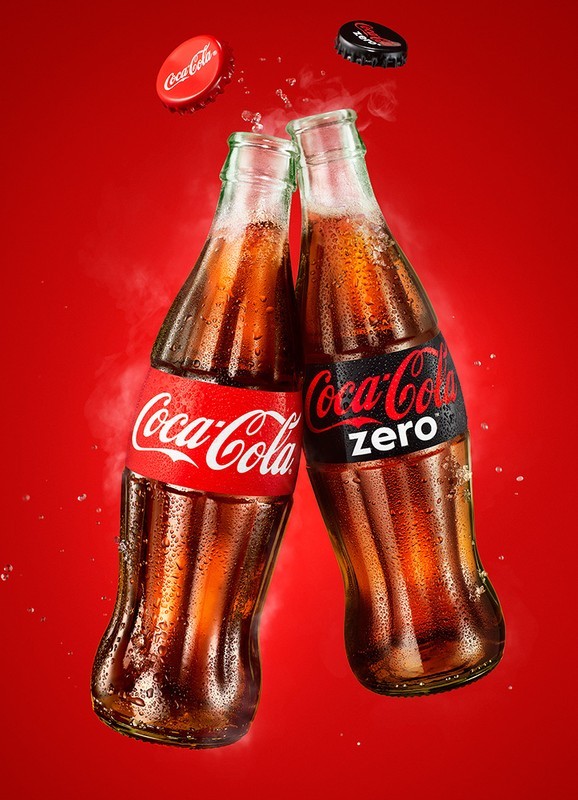 Two glass Coca-Cola bottles touch in a toast against a solid red backdrop with floating caps. The bottles feature the Coca-Cola logo.