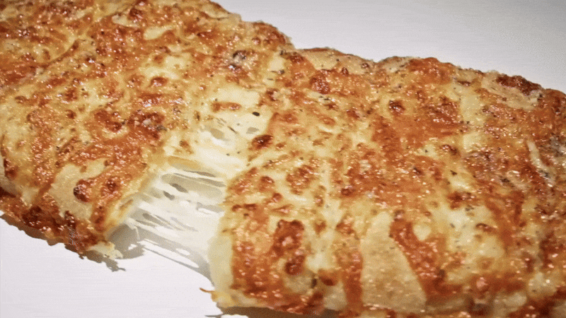 Close-up of a cheese-covered pastry being pulled apart, revealing stringy, melted cheese. The pastry has a golden-brown, bubbly crust.