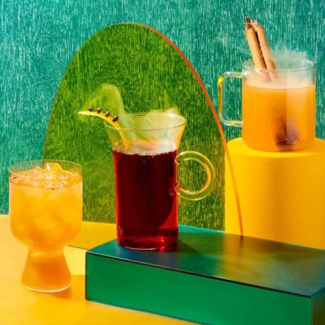 Three glass mugs with colorful drinks, garnished and arranged on geometric platforms against a patterned backdrop for a vibrant food still life.
