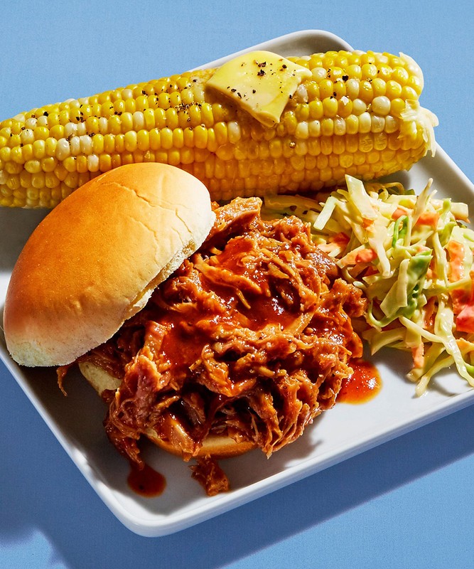 Overhead shot on a blue background: pulled pork sandwich, corn on the cob with butter, and coleslaw on a white square plate.