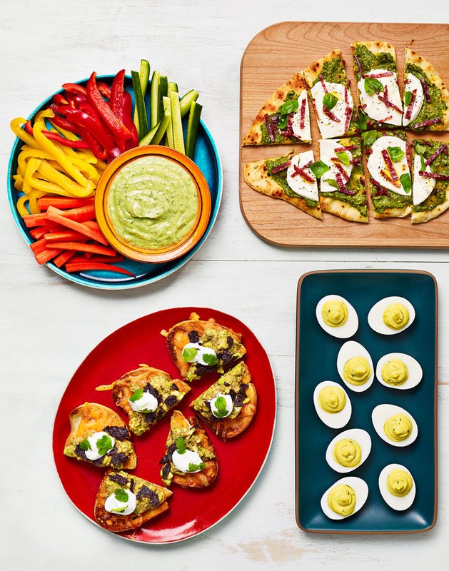 Overhead shot of appetizer spread: vegetable sticks with dip, sliced pizza on wood, bruschetta on a red plate, and deviled eggs.