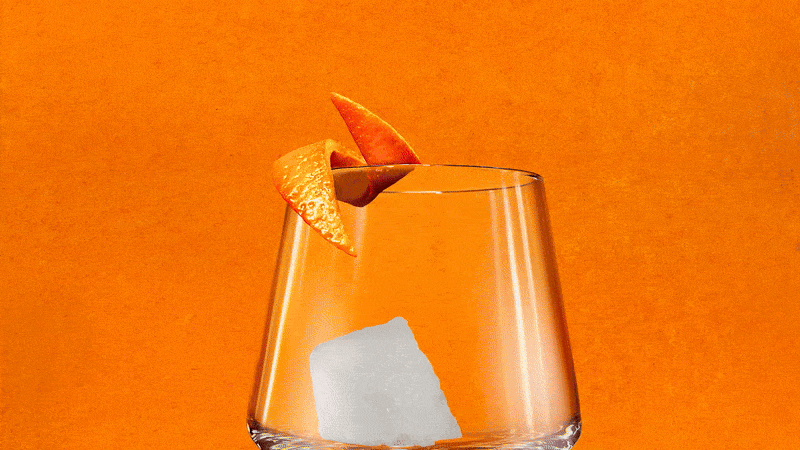 Close-up of a cocktail glass with an ice cube, an orange peel garnish, and a textured orange background.