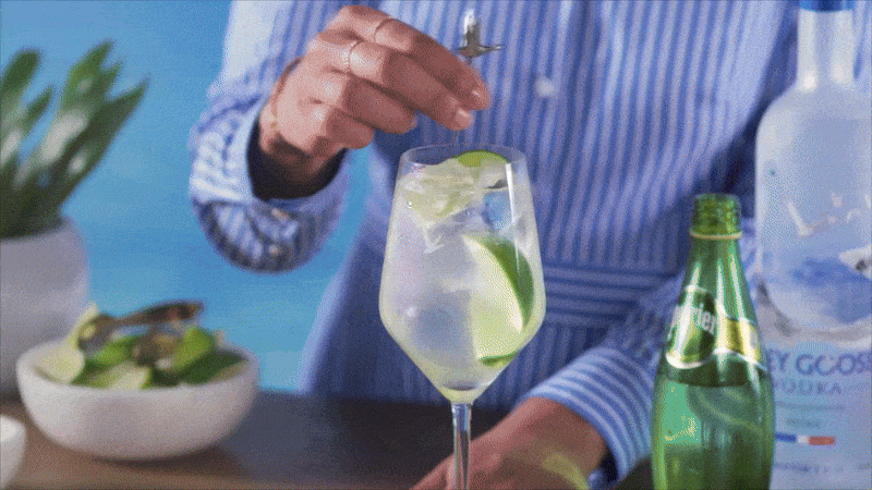 Person's hand adding a garnish to a cocktail in a tall glass, with bottles, a dish of lime, and plant in the background.