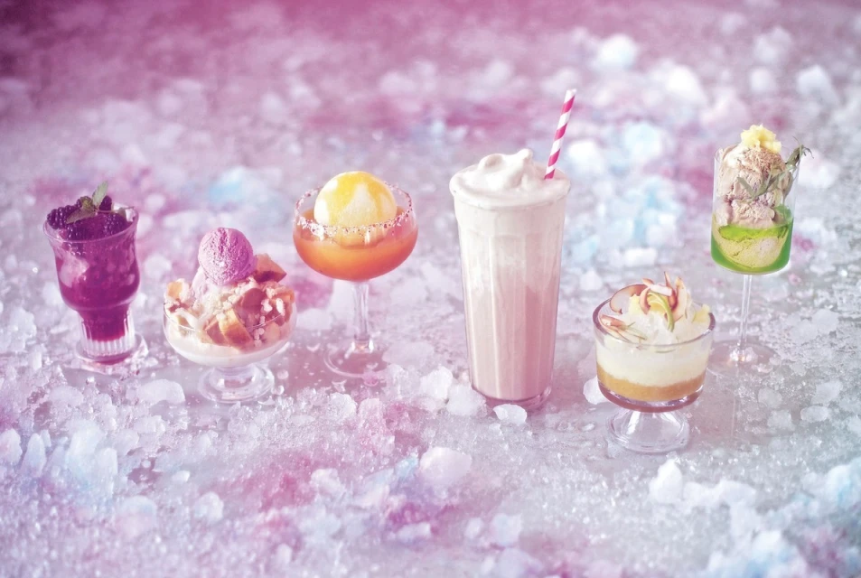 Several dessert beverages are lined up on a bed of crushed ice, with a pink and blue gradient background.