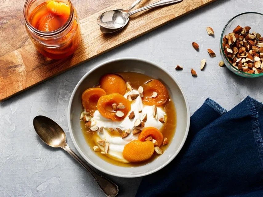 Overhead shot of a bowl of apricots and yogurt, with a jar of apricots, spoons, almonds, and a blue cloth on a grey surface.
