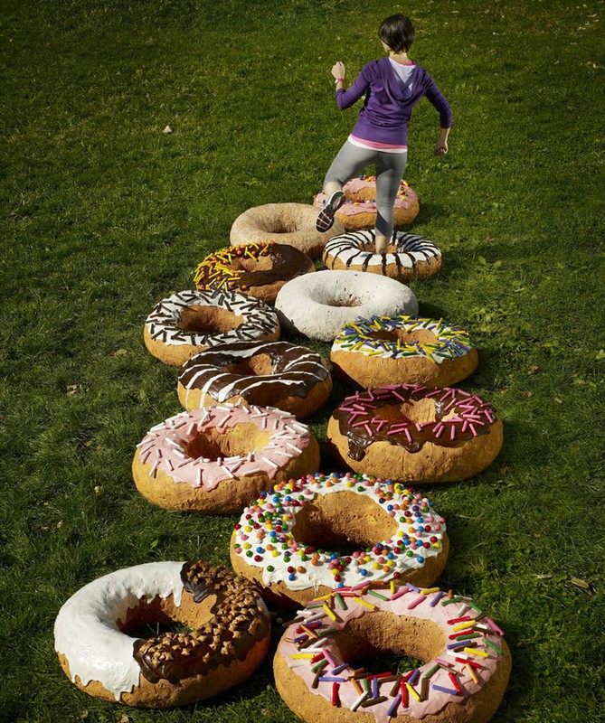 A woman steps onto a row of giant novelty donuts placed on a grassy lawn outdoors in daylight for a fitness or food and beverage theme.