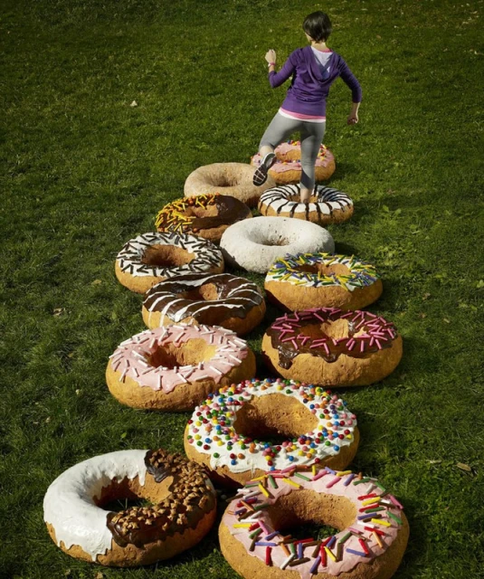 A woman steps onto a row of giant novelty donuts placed on a grassy lawn outdoors in daylight for a fitness or food and beverage theme.