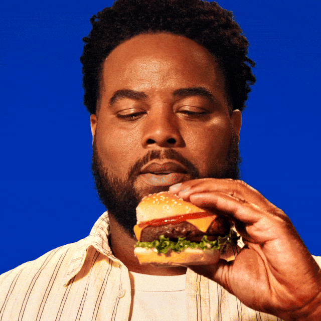 An adult male with dark hair and a beard holds a cheeseburger in front of a blue background, posing as if to take a bite.