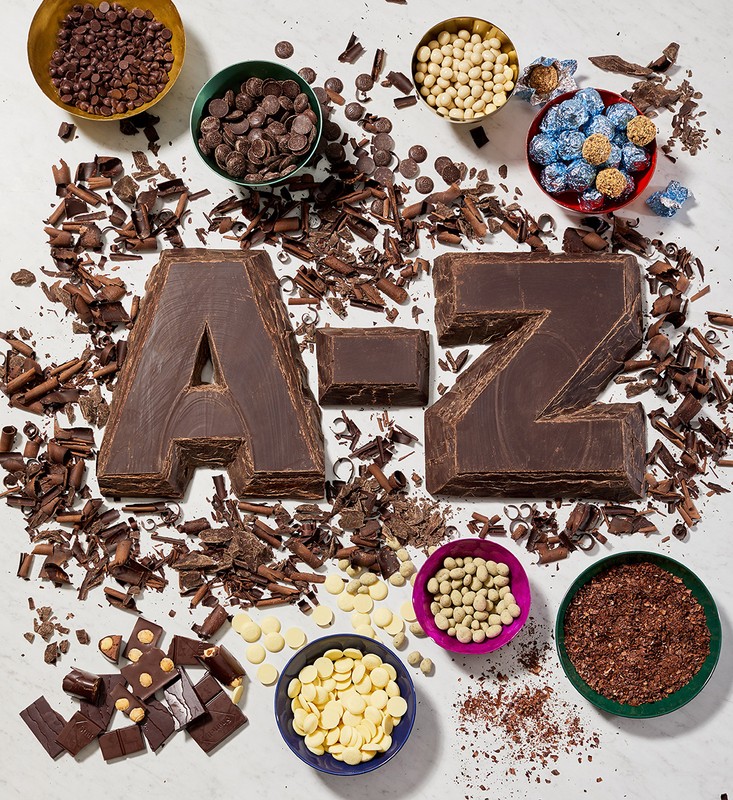 Overhead shot of chocolate letters spelling 'A-Z' surrounded by bowls and piles of chocolate chips, shavings, and candies on white surface.