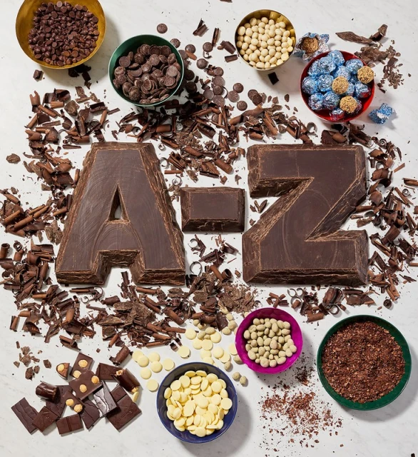 Overhead shot of chocolate letters spelling 'A-Z' surrounded by bowls and piles of chocolate chips, shavings, and candies on white surface.