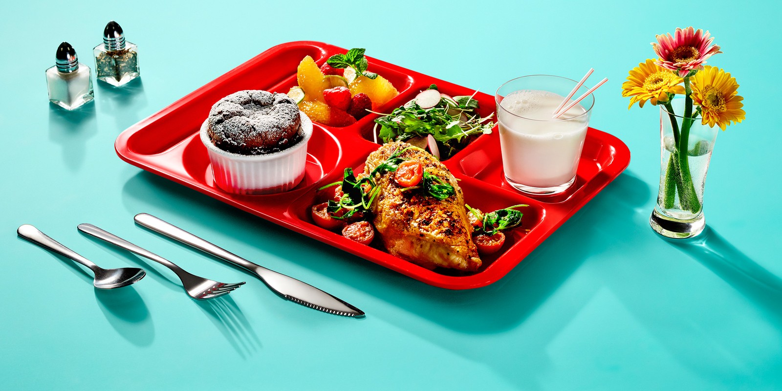 Overhead shot of a school lunch tray with a chicken breast, salad, fruit, muffin, milk, utensils, and flowers against a blue background.