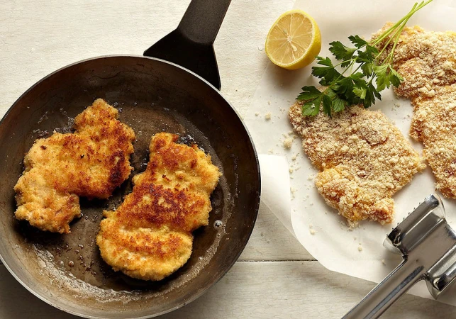 Overhead shot of breaded cutlets in a pan and on parchment paper with a lemon half and meat tenderizer for food photography.
