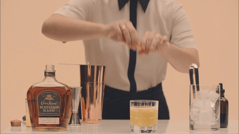Bartender squeezing orange into cocktail over white table with copper shaker, glasses, whiskey bottle, ice. Cream and black outfit.