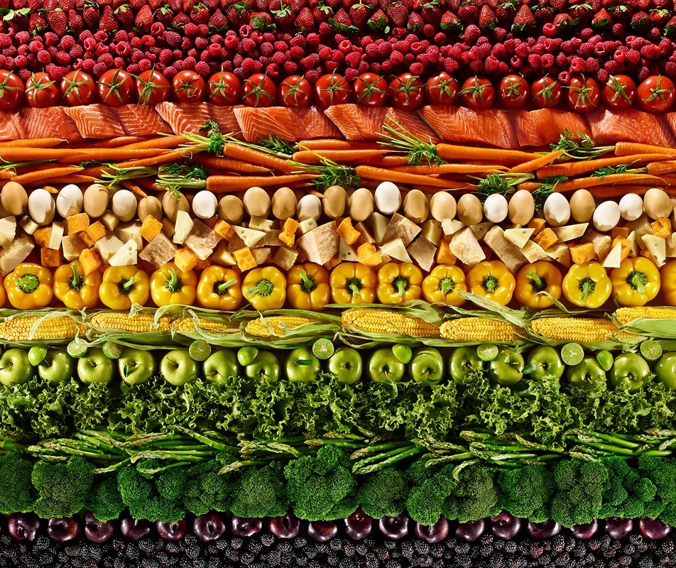 Overhead shot of rows of colourful fruits and vegetables, including berries, tomatoes, salmon, carrots, eggs, cheese, corn, apples, and lettuce.