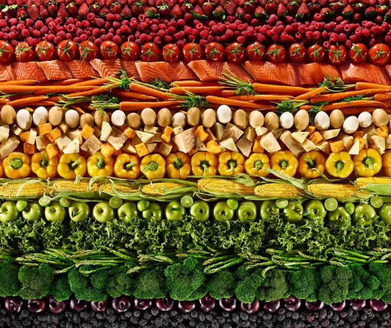 Overhead shot of rows of colourful fruits and vegetables, including berries, tomatoes, salmon, carrots, eggs, cheese, corn, apples, and lettuce.