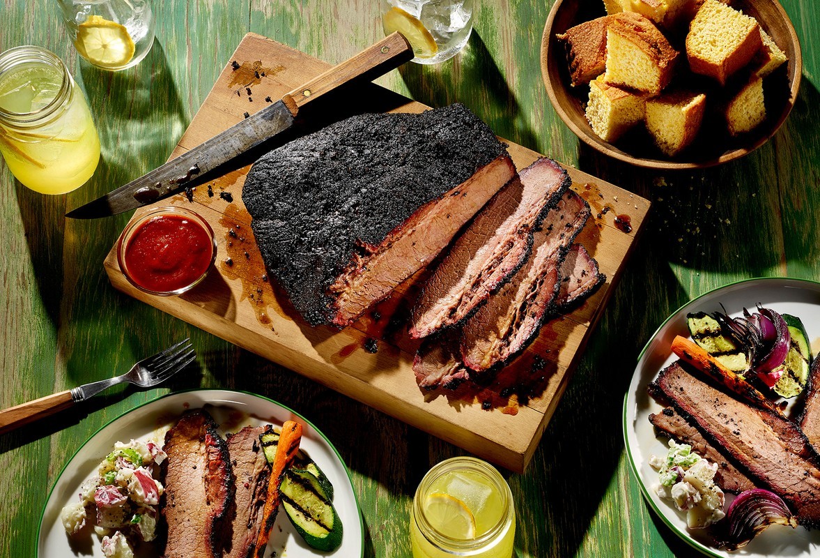 Overhead shot of sliced brisket on a wooden board with sides of potato salad, grilled vegetables, cornbread, lemonade, and ketchup.