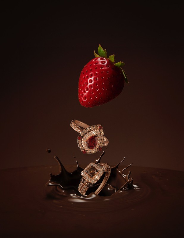 A strawberry and two diamond rings falling into a splash of chocolate on a brown background in a high-angle product shot.